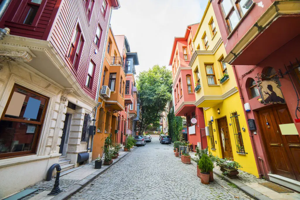 İstanbul'un tarihi mahallelerinden birinde yer alan Arnavut kaldırımlı dar bir sokak, restore edilmiş renkli Osmanlı evleri, ahşap pencereler ve bina girişlerindeki saksı bitkileriyle birlikte görülmektedir. Sokağın sonunda yükselen yeşil ağaçlar ve sakin atmosfer, İstanbul'un tarihi konut dokusunu ve kültürel kimliğini yansıtmaktadır.