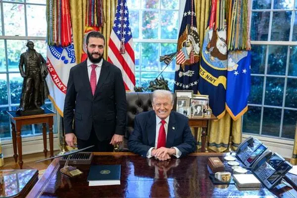 ABD Başkanı Donald Trump, Beyaz Saray'daki Oval Ofis'te başkanlık masasının arkasında otururken, Suriye Geçiş Hükümeti Başkanı Ahmed eş-Şara koyu renk takım elbise ve kırmızı kravatıyla yanında ayakta duruyor. Arka planda ABD bayrakları, askerî kuvvetlere ait sancaklar ve Oval Ofis'in büyük pencereleri görülüyor.