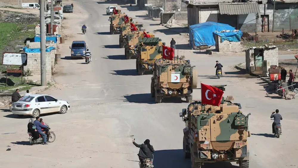 Suriye’nin kuzeyindeki kentsel bölgelerden birinde ilerleyen Türk askeri zırhlı araç konvoyu. Türk bayrakları taşıyan zırhlı araçlar, sivil halkın günlük hareketliliği arasında bir cadde üzerinden ilerlerken görülüyor.
