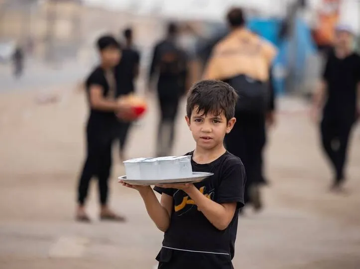 Erbain yürüyüşü güzergâhındaki Iraklı bir çocuk, ziyaretçilere ikram etmek üzere su bardaklarıyla dolu bir tepsi taşıyor; Irak halkının misafirperverlik kültüründe hizmetin, cömertliğin ve çocukların katılımının anlamlı bir örneği.