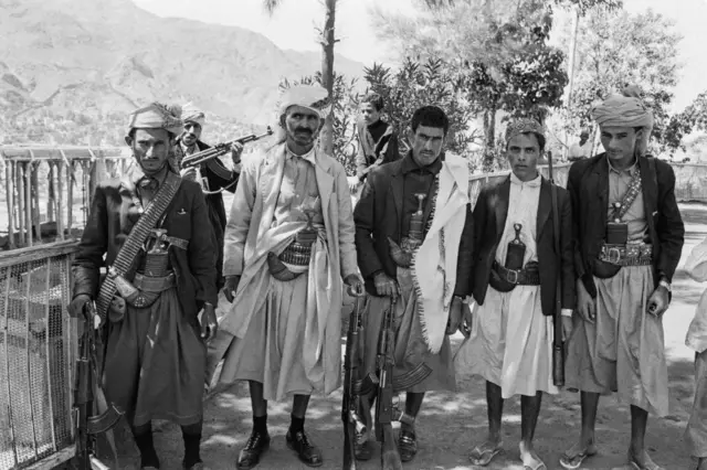 Dağlık bir bölgede, geleneksel kıyafetleri, cenbiye hançerleri ve hafif silahlarıyla görülen bir grup Yemenli erkeğin siyah-beyaz fotoğrafı. Kültürel ve dinî faaliyetlerle ortaya çıkan, daha sonra Husi hareketi ile Ensarullah’ın oluşumuna zemin hazırlayan “Mümin Gençlik” hareketini simgelemektedir.