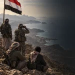 Kızıldeniz’e hâkim yüksek bir noktadaki Yemenli savaşçıları, Yemen bayrağını ve Hudeyde’den Muha ile Babülmendep yakınındaki Meyyun Adası’na uzanan ilerleme güzergâhını gösteren haber illüstrasyonu.
