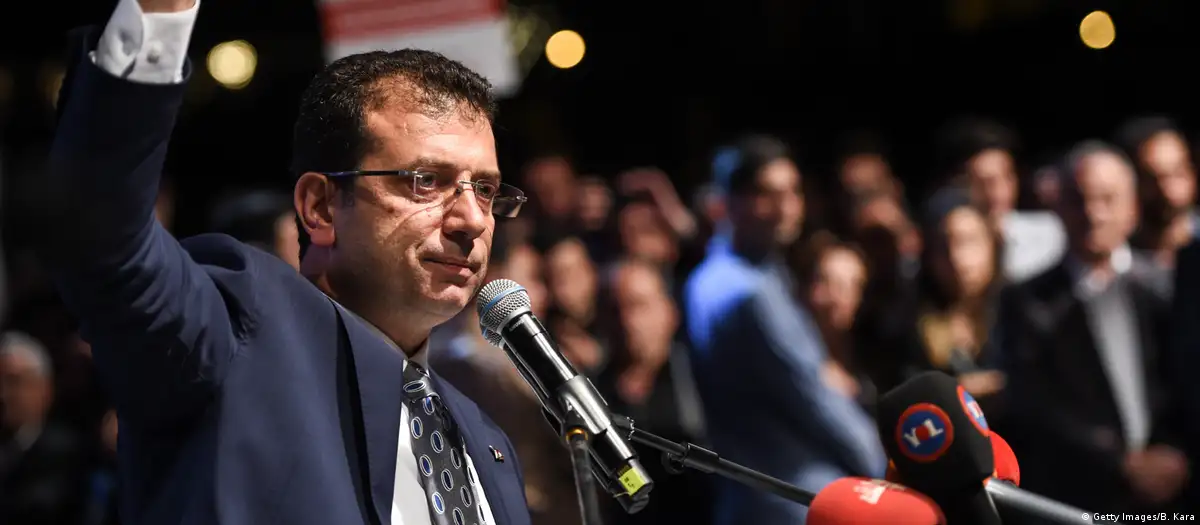 Lacivert takım elbiseli ve gözlüklü Ekrem İmamoğlu, arkasındaki kalabalığın önünde bir elini kaldırarak mikrofonda konuşuyor.