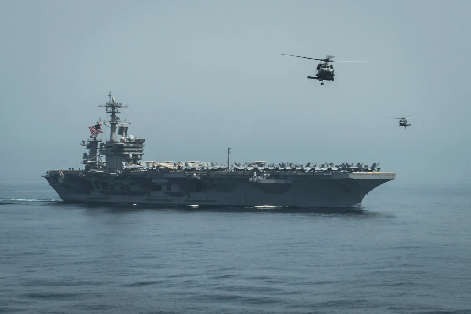 Güvertesinde savaş uçakları bulunan ve üzerinde iki helikopterin uçtuğu Amerikan uçak gemisi; İran çevresindeki artan ABD askerî varlığını yansıtan bir görüntü.