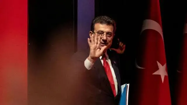Koyu renk takım elbiseli ve kırmızı kravatlı Ekrem İmamoğlu, Türk bayrağının yanında bir elinde dosya tutarken diğer elini selamlamak için kaldırıyor.